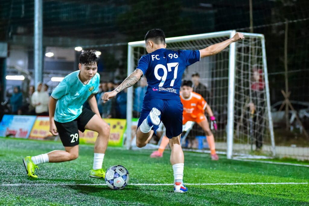 Blue attacker lines up a shot as defender and goalkeeper react.